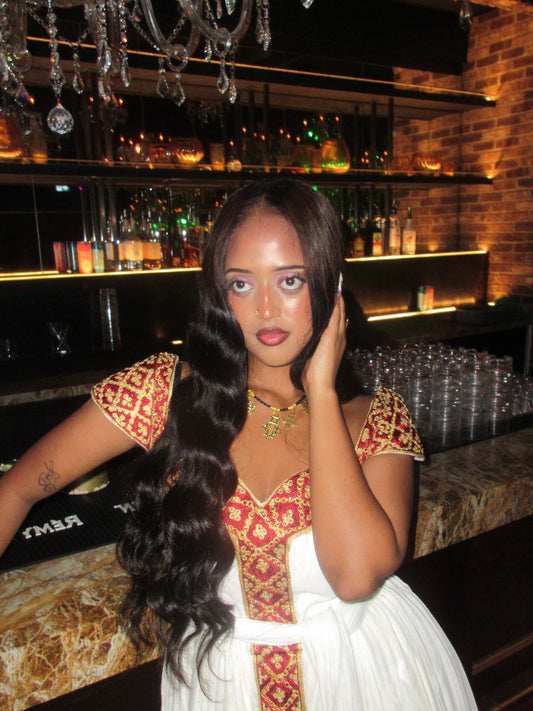 A woman posing in a white dress with red and gold embroidered Habesha Kemis, set against a dimly lit bar background.