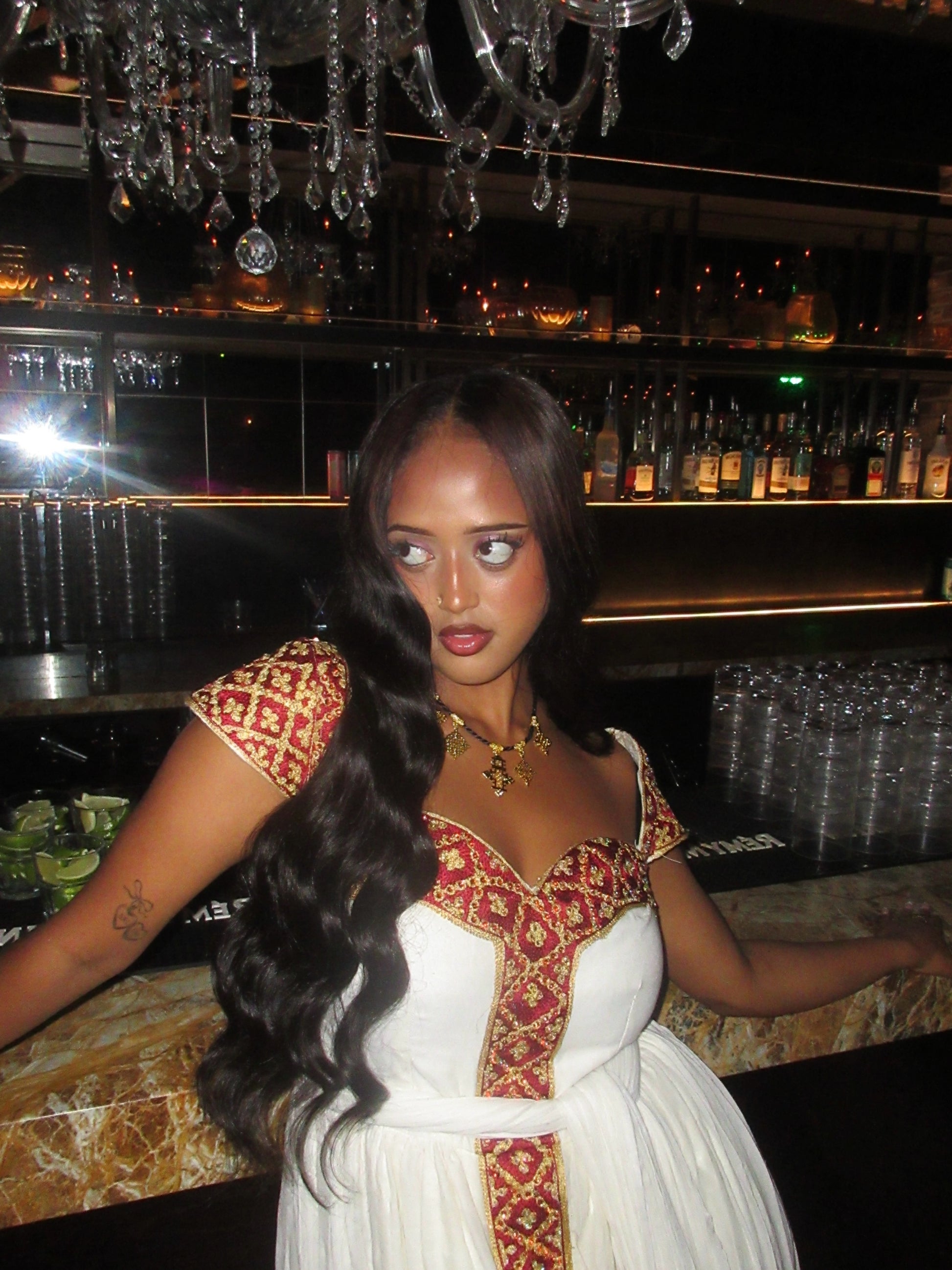 A woman with long, wavy black hair stands behind a bar counter wearing a white Habesha Kemis with ornate red and gold embroidery. The background shows a dimly lit bar interior with shelves of bottles, glasses, and a chandelier.