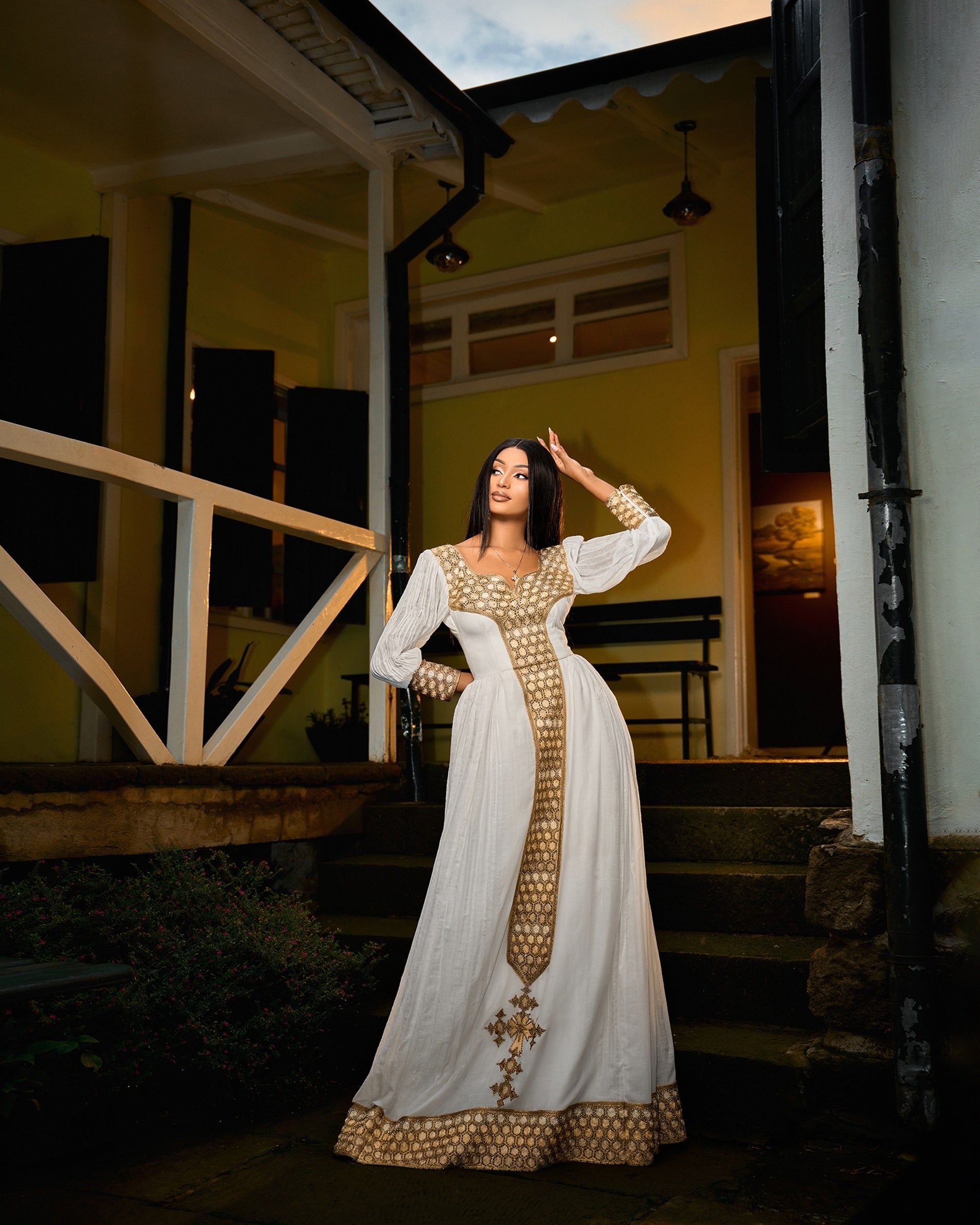 A woman wearing a long Habesha Kemis with gold patterned trim and a gold cross detail on the front, posing on the steps of a house at dusk.