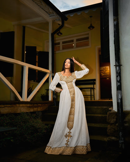 A woman wearing a long Habesha Kemis with gold patterned trim and a gold cross detail on the front, posing on the steps of a house at dusk.