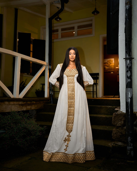 A woman stands on outdoor steps at night wearing a long gold Habesha Kemis with a gold patterned front panel, sleeves, and hem. The background shows a dimly lit porch with yellow walls.