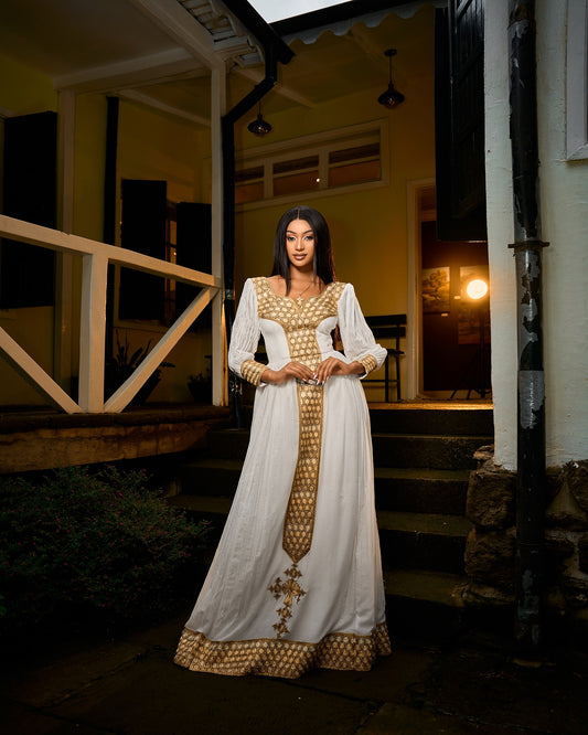 A woman wearing a long white traditional Habesha Kemis with gold geometric patterns and sheer sleeves, standing on stone steps at night in front of a house.