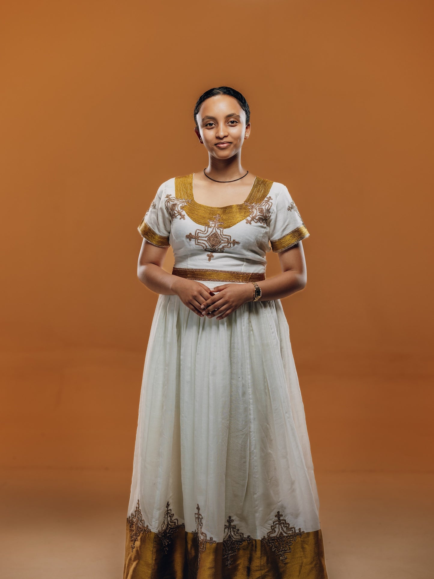 A woman modeling a traditional white and gold embroidered habesha kemis  against a solid warm brown studio background.