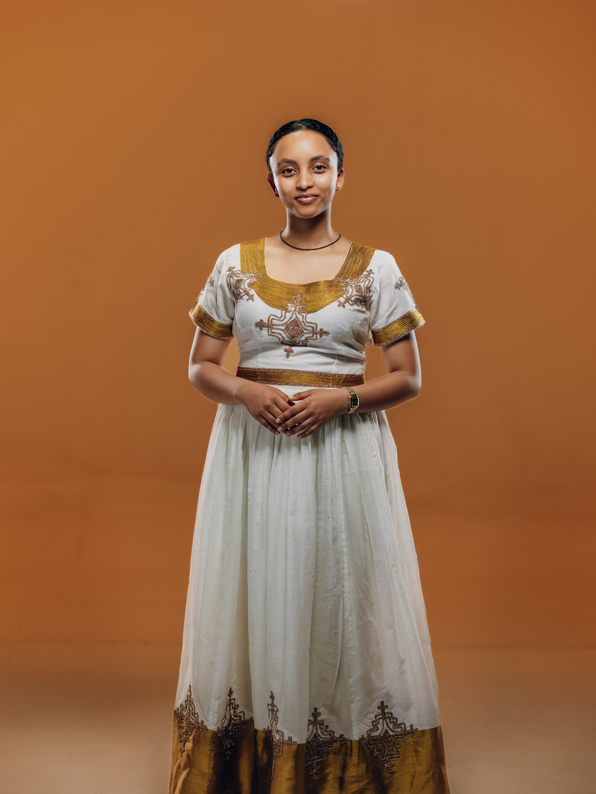 A woman modeling a traditional white and gold embroidered habesha kemis  against a solid warm brown studio background.