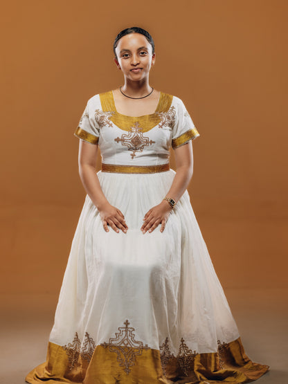 A woman wearing a traditional Ethiopian white and gold habesha kemis  with ornate embroidery, standing against a solid brown studio background.