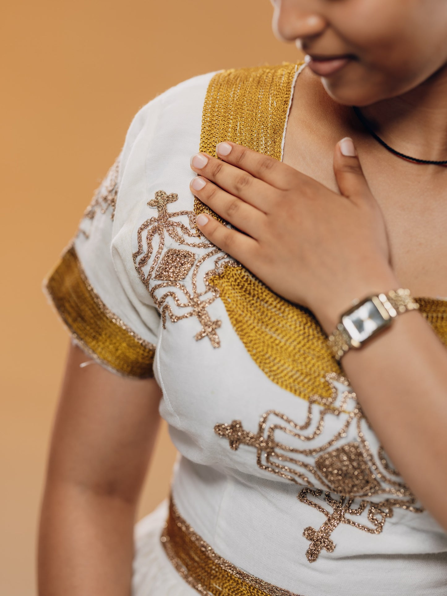 A close-up of a person wearing a white embroidered habesha kemis  with gold metallic embroidery and trim, against a warm beige background.