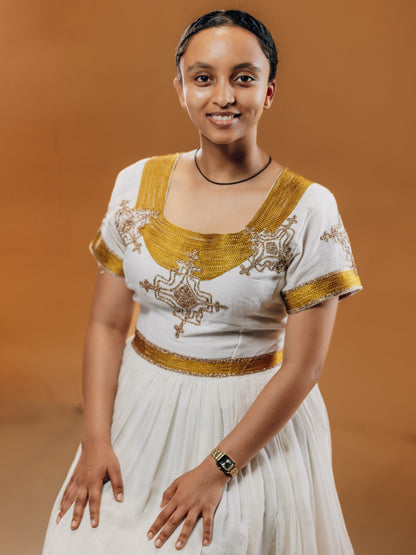 A woman wearing a traditional habesha kemis with gold embroidery and a gold waistband, standing against a solid brown studio background.