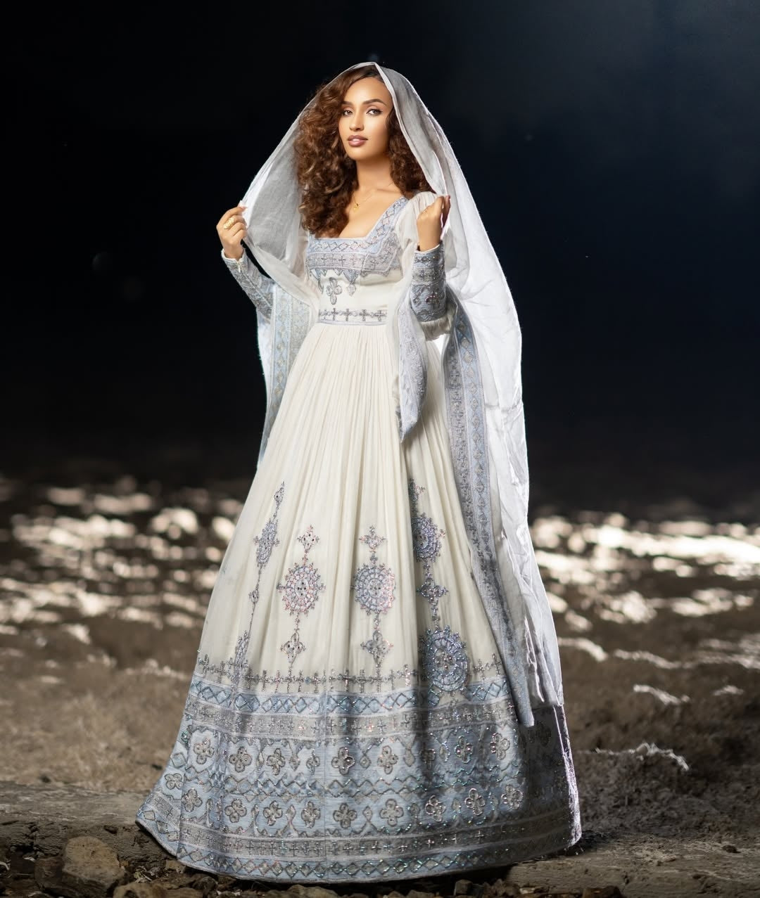 Woman in a white and blue habesha traditional outfit with a veil against a dark background.