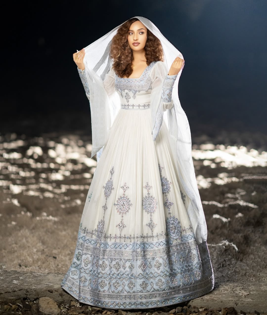 Woman in a white and blue habesha traditional dress standing on a rocky shore with water in the background.