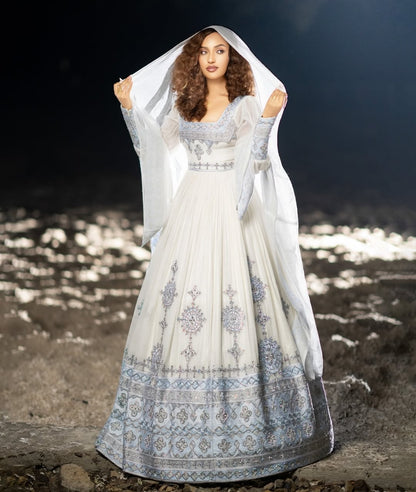 Woman in a white and blue habesha traditional dress standing on a rocky shore with water in the background.