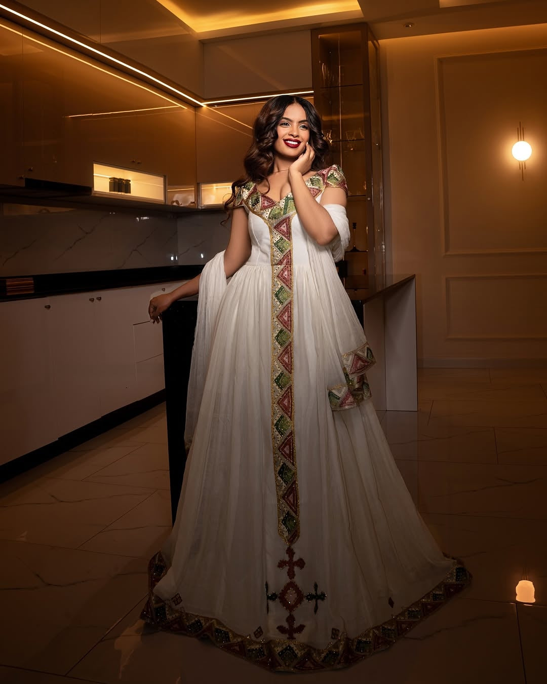 Woman in a white habesha dress with colorful embroidery standing indoors.