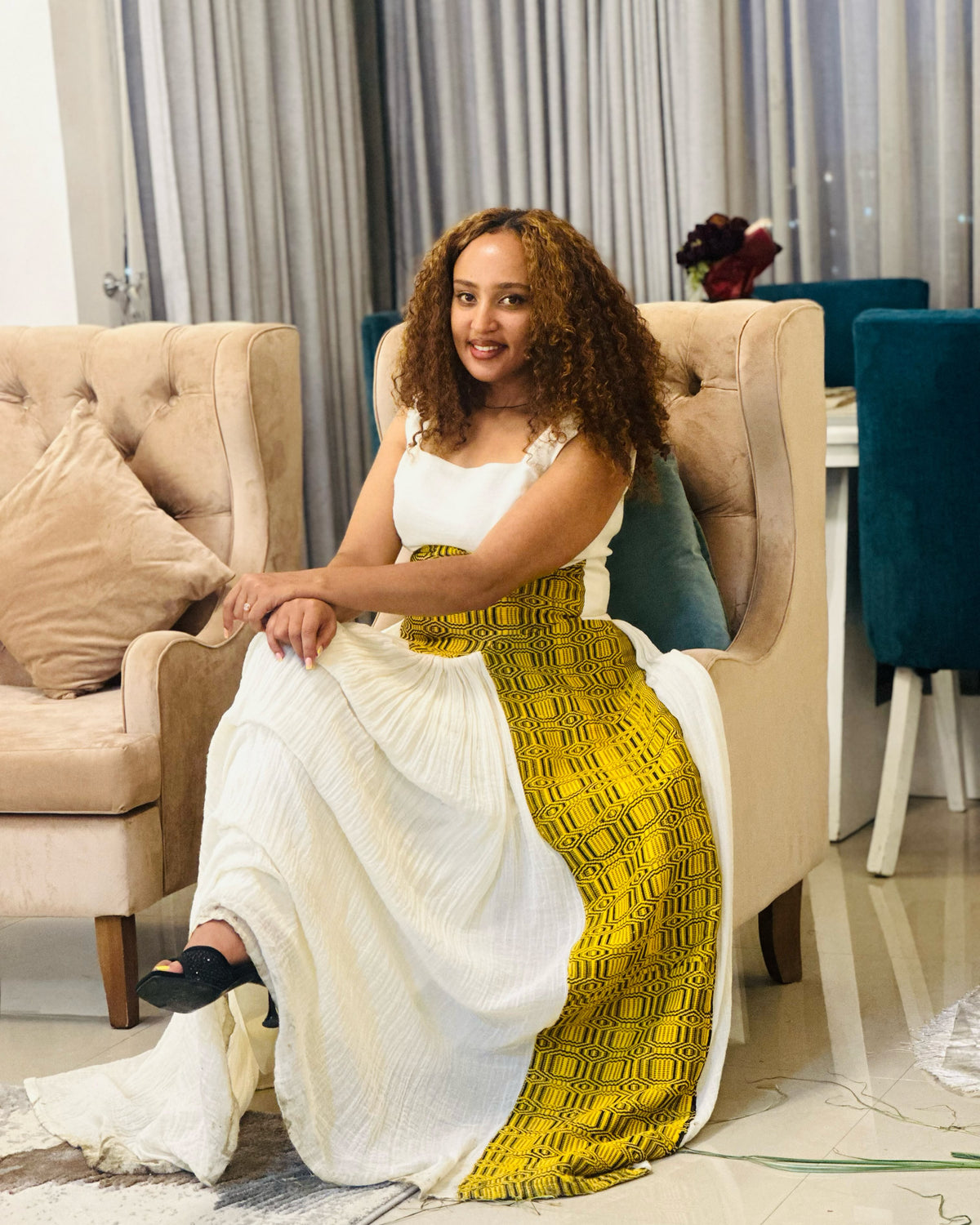 A woman with curly hair sits in a beige armchair wearing a sleeveless white Ethiopia store dress with a wide yellow patterned side panel, in a modern living room setting.