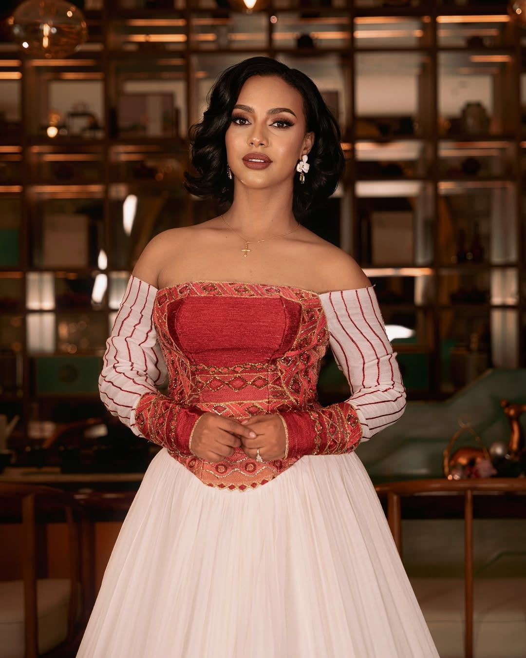 Woman wearing a white Ethiopian cultural dress with red patterned hem against a plain background