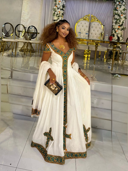 A woman wearing a white traditional Habesha Kemis with green and gold embroidery, holding a black clutch bag, standing in a decorated event hall with white steps and gold accents.