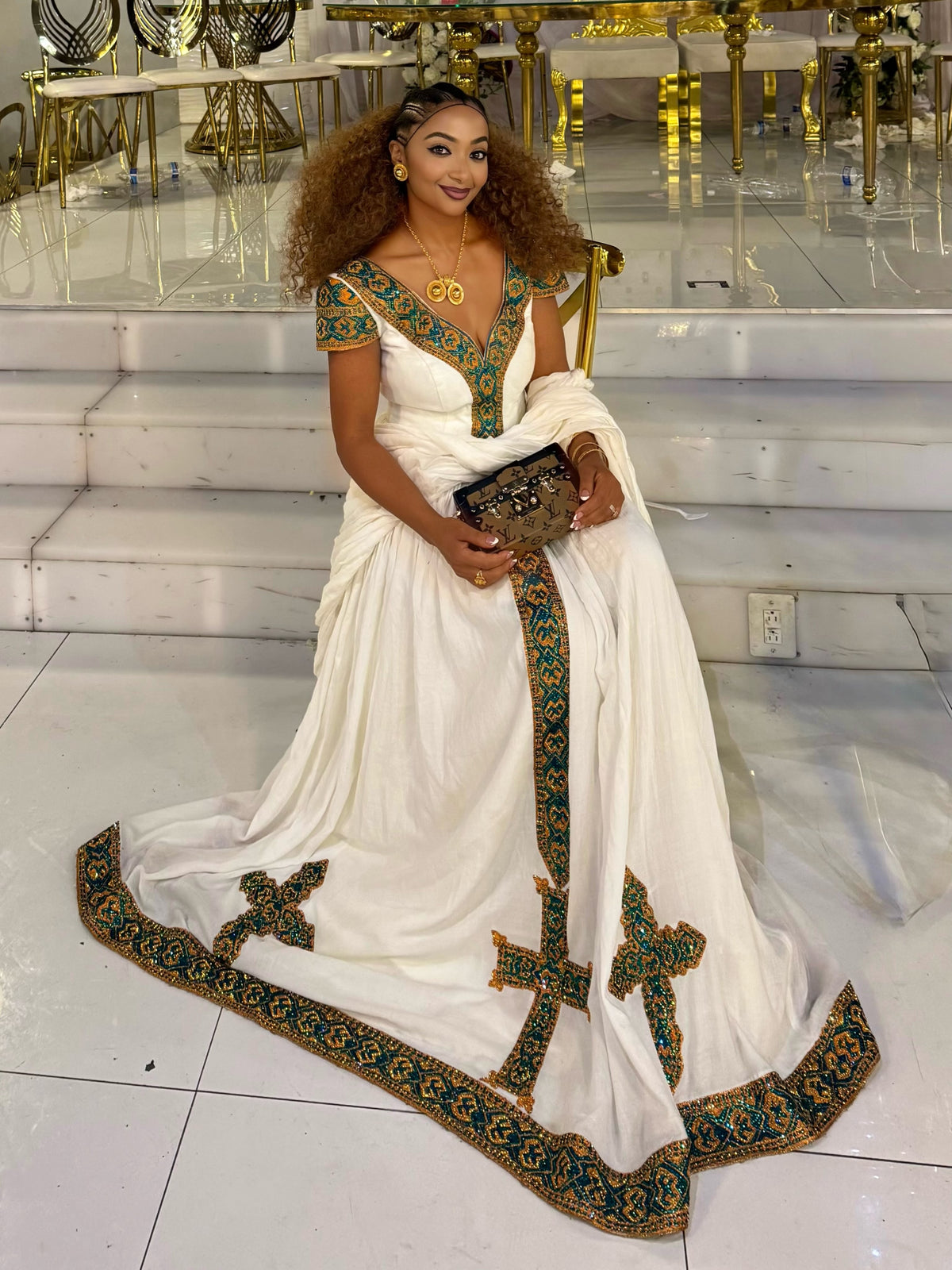 A woman wearing a white traditional Habesha Kemis with ornate green and gold embroidered borders and a matching embroidered blouse, seated on white steps in a decorated interior.