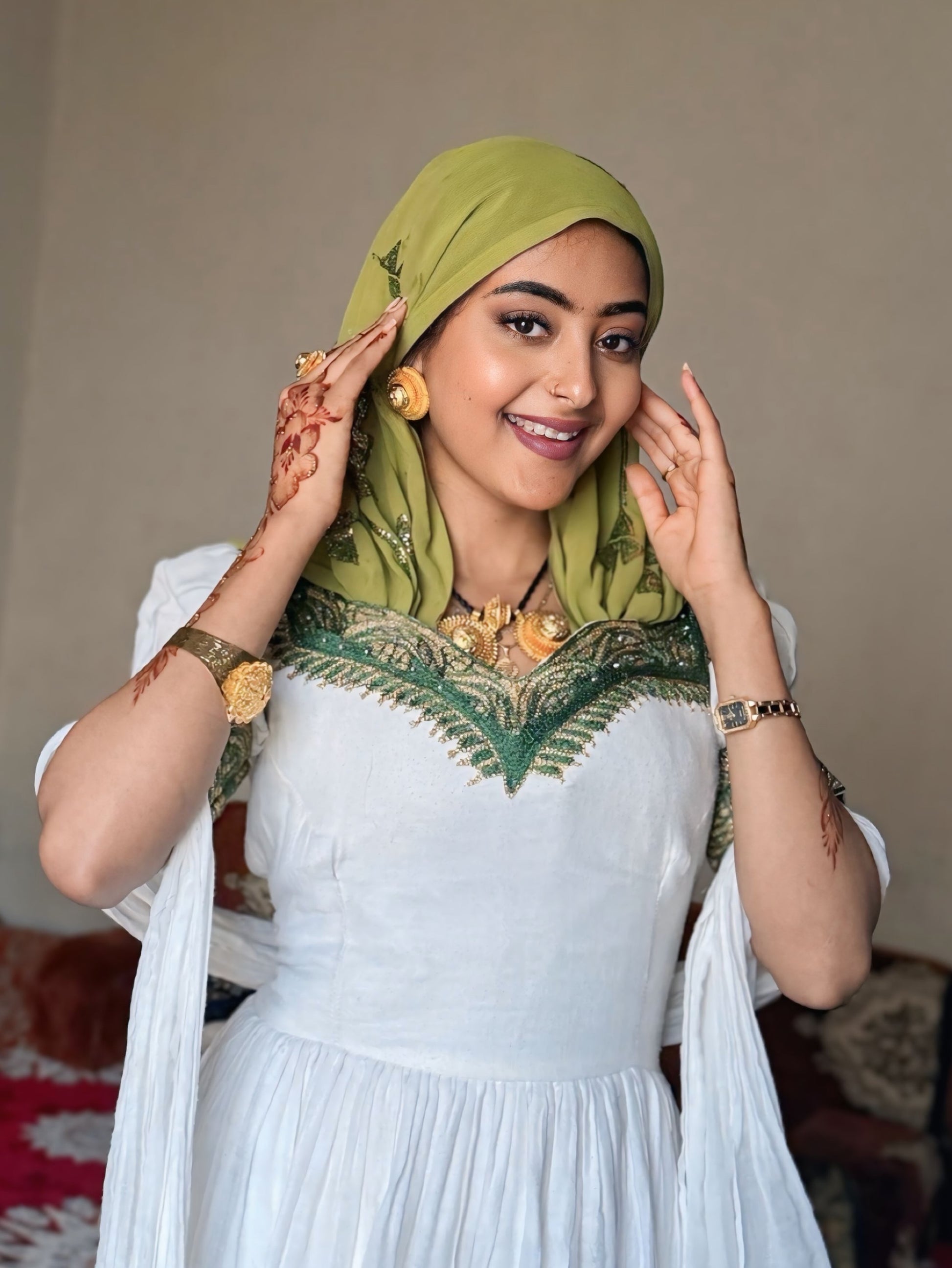 A woman wearing a white  Habesha Dress  with green embroidered detailing and a light olive green hijab, posing indoors with her hands near her head.