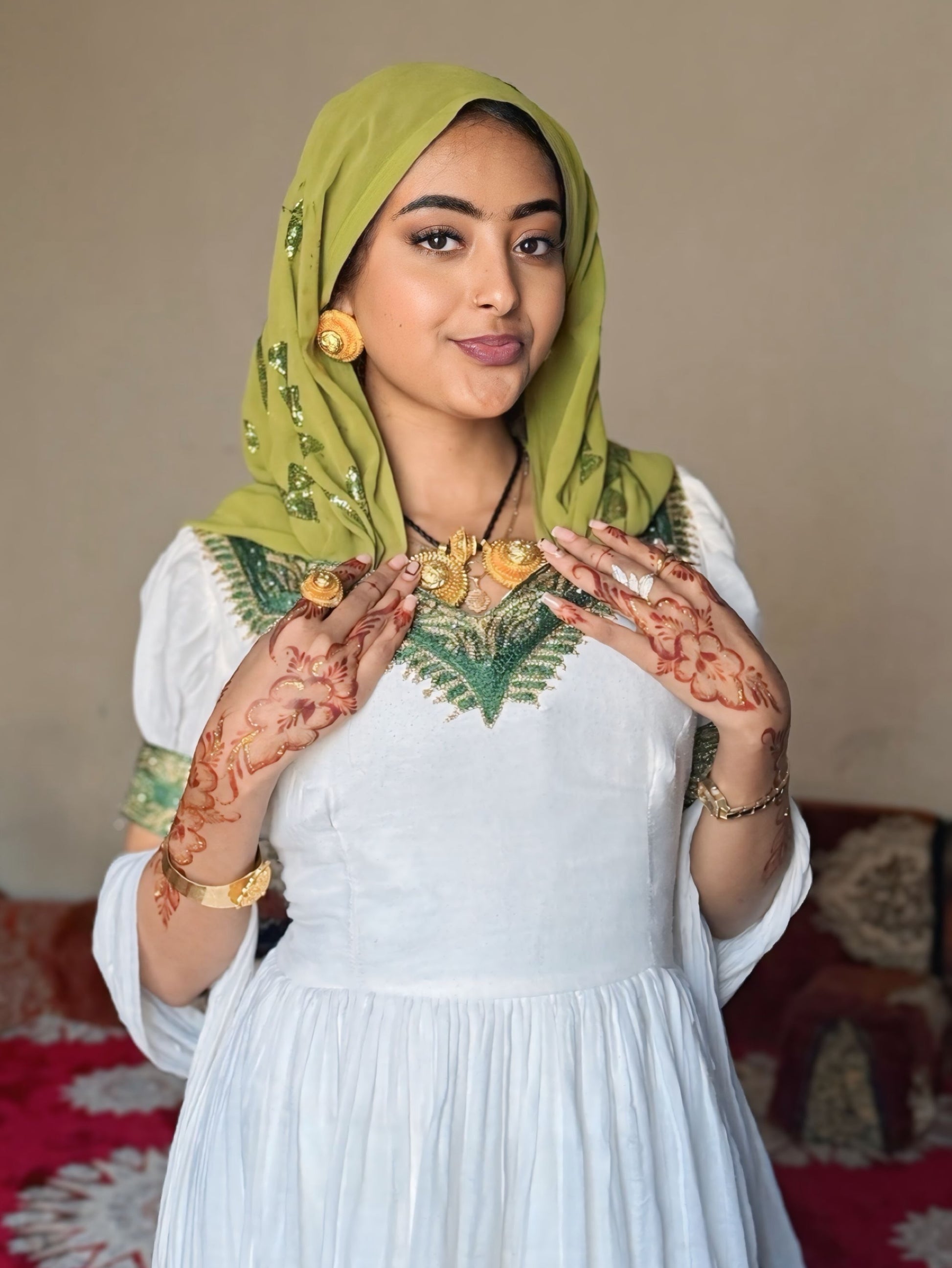 A woman wearing a white traditional  Habesha Dress  with green embroidered details, a green hijab, and gold jewelry, posing with her hands decorated with henna.