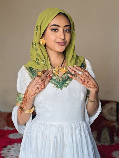 A woman wearing a white traditional  Habesha Dress  with green embroidered details, a green hijab, and gold jewelry, posing with her hands decorated with henna.
