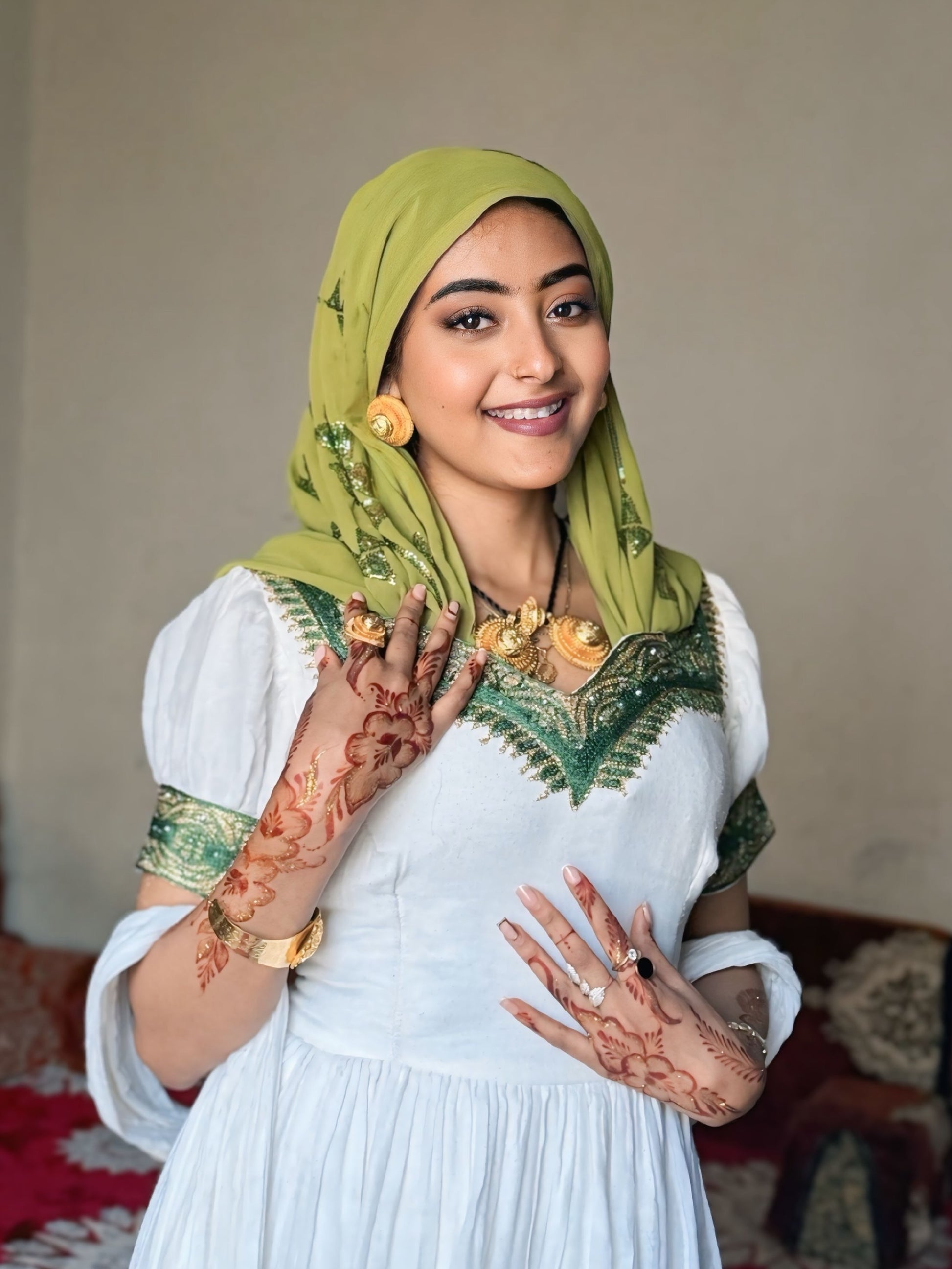 A woman wearing a white traditional  Habesha Dress  with green embroidered details, a green patterned hijab, and gold jewelry, posing indoors with henna on her hands.