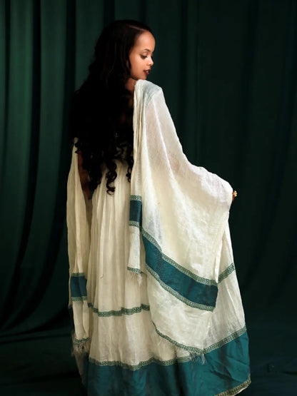 A woman wearing a white and teal traditional habesha kemis, shown from the back against a dark green curtain backdrop.