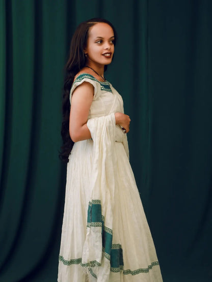 A woman wearing a traditional white habesha kemis with green border embroidery, standing against a dark green draped backdrop.