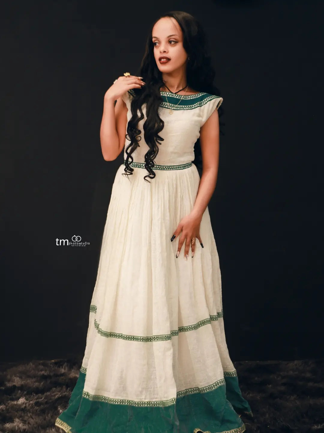 A woman modeling a long cream-colored dress with green habesha kemis and a green hem, set against a dark studio background.