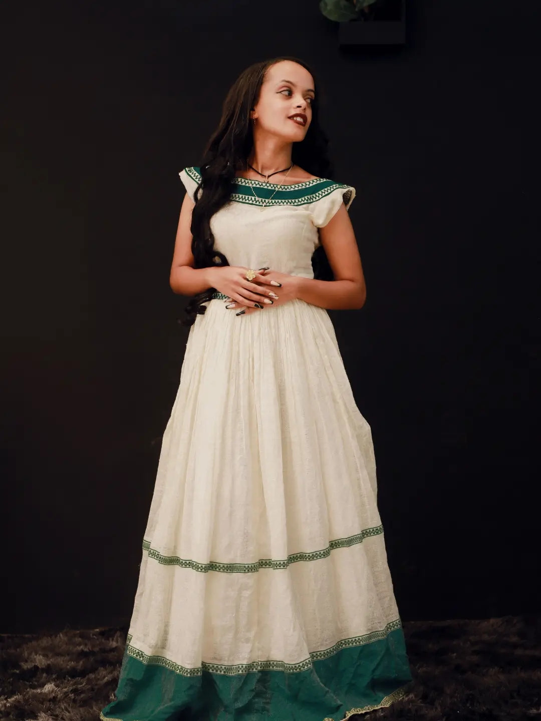 A woman wearing a long cream-colored habesha kemis with green border detailing, standing against a dark background.