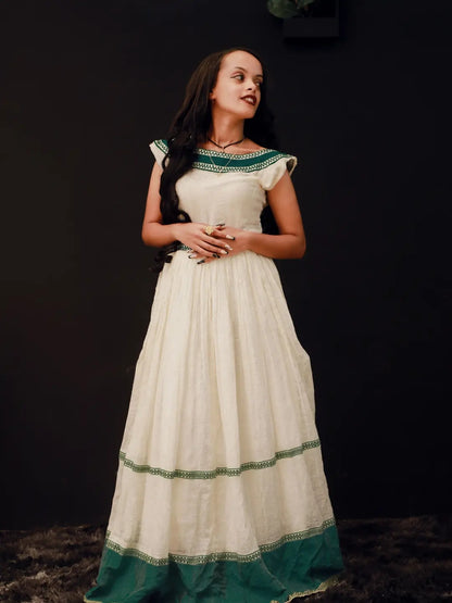 A woman wearing a long cream-colored habesha kemis with green border detailing, standing against a dark background.
